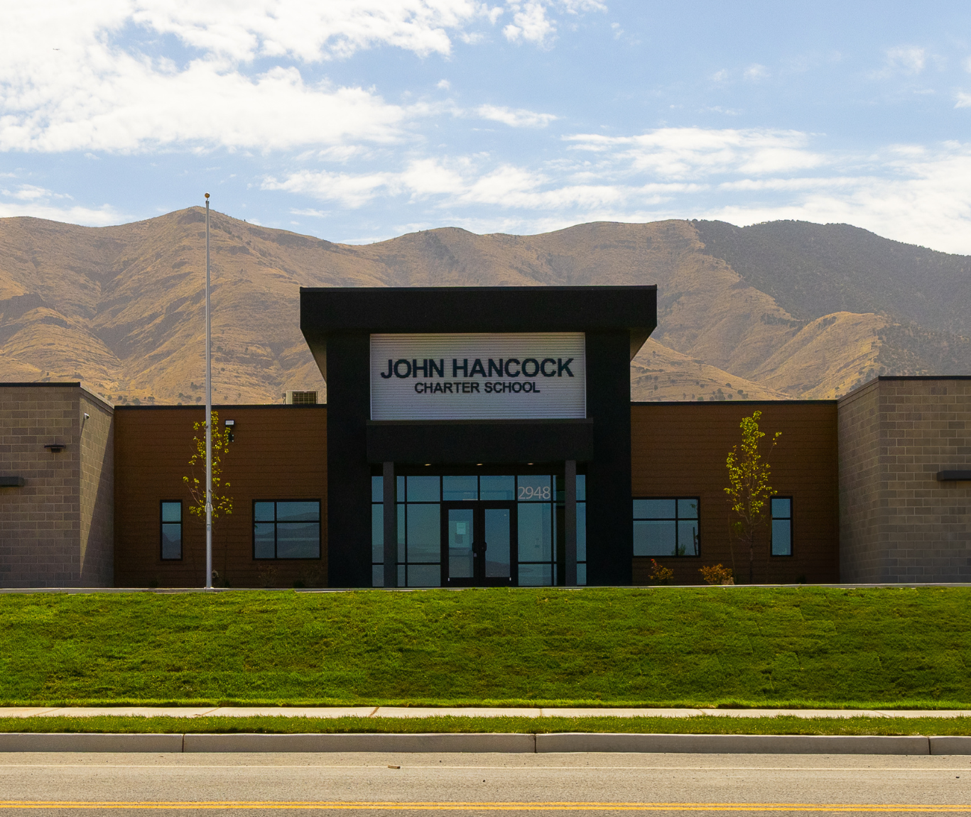 A modern school building, John Hancock Charter School, with mountains rising in the background under a blue sky.